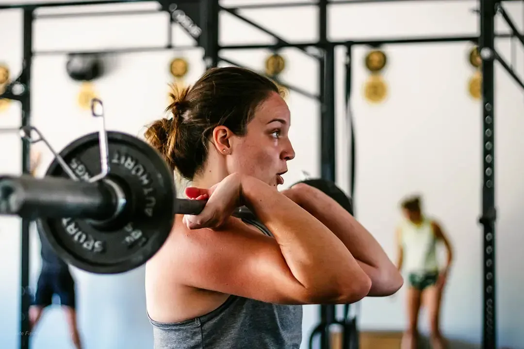 Woman in gym holding barbell, preparing to lift. Gray tank top, focused expression. Other people and equipment in background.