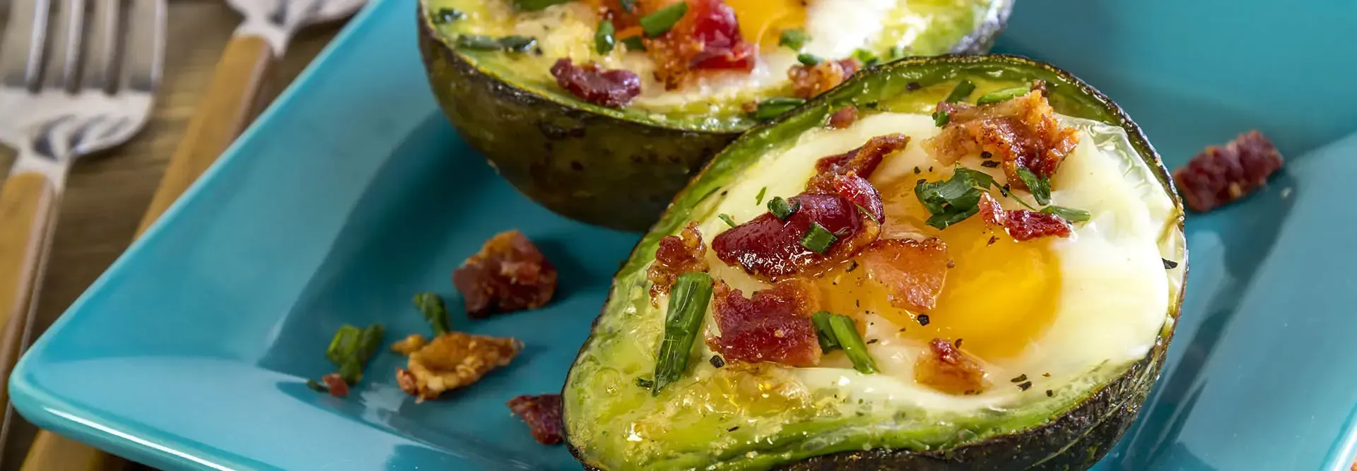 Baked eggs in avocado halves with bacon bits on a turquoise plate.