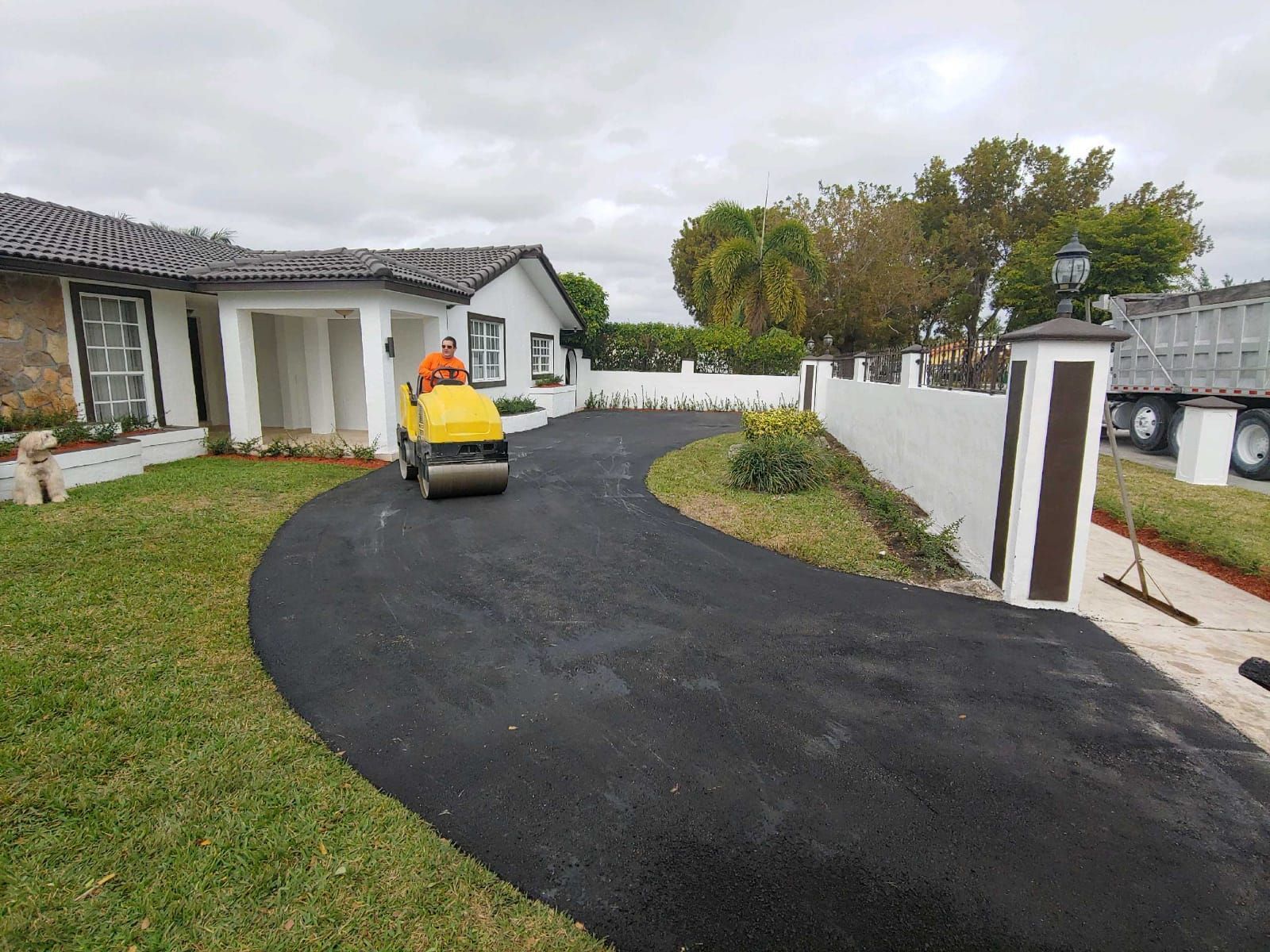 A yellow roller is rolling asphalt in a driveway in front of a house.