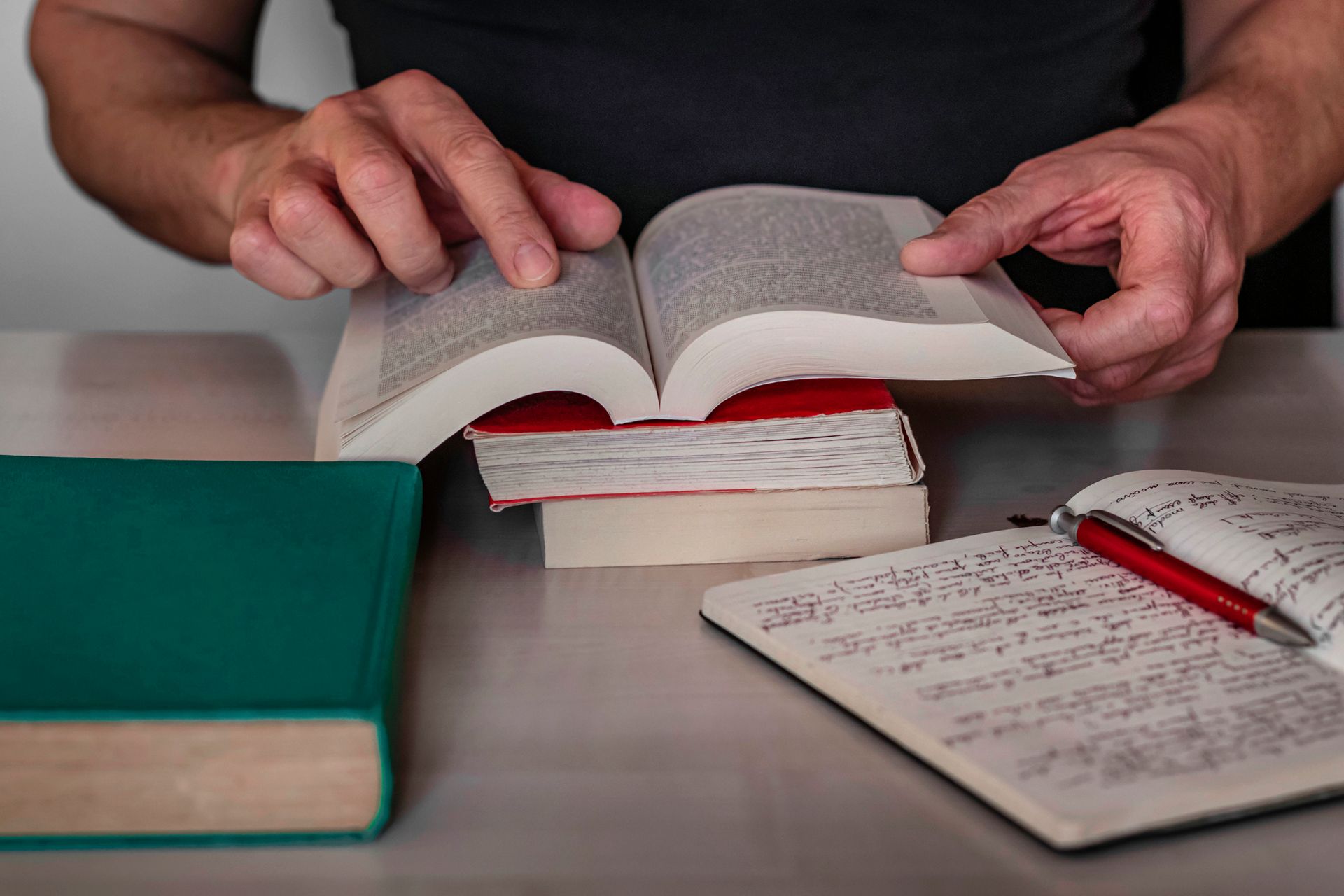 Persona che legge un libro, aperto su una pila di libri, con un quaderno e una penna su un tavolo.