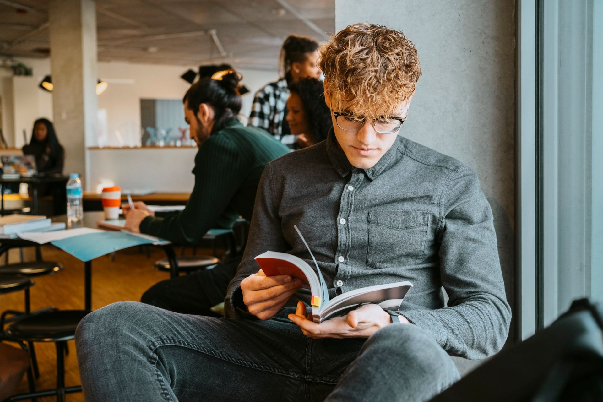 Un giovane con gli occhiali legge un libro, seduto vicino alla finestra di un bar.
