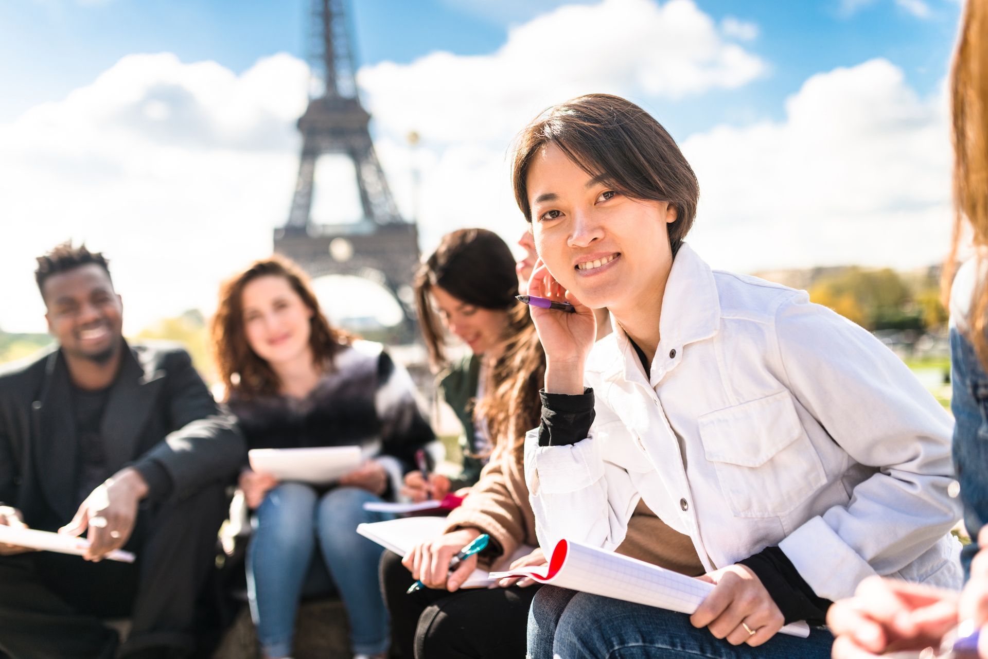 Un gruppo di studenti davanti alla Torre Eiffel, sorridenti mentre lavorano ai loro elaborati. Giornata di sole.