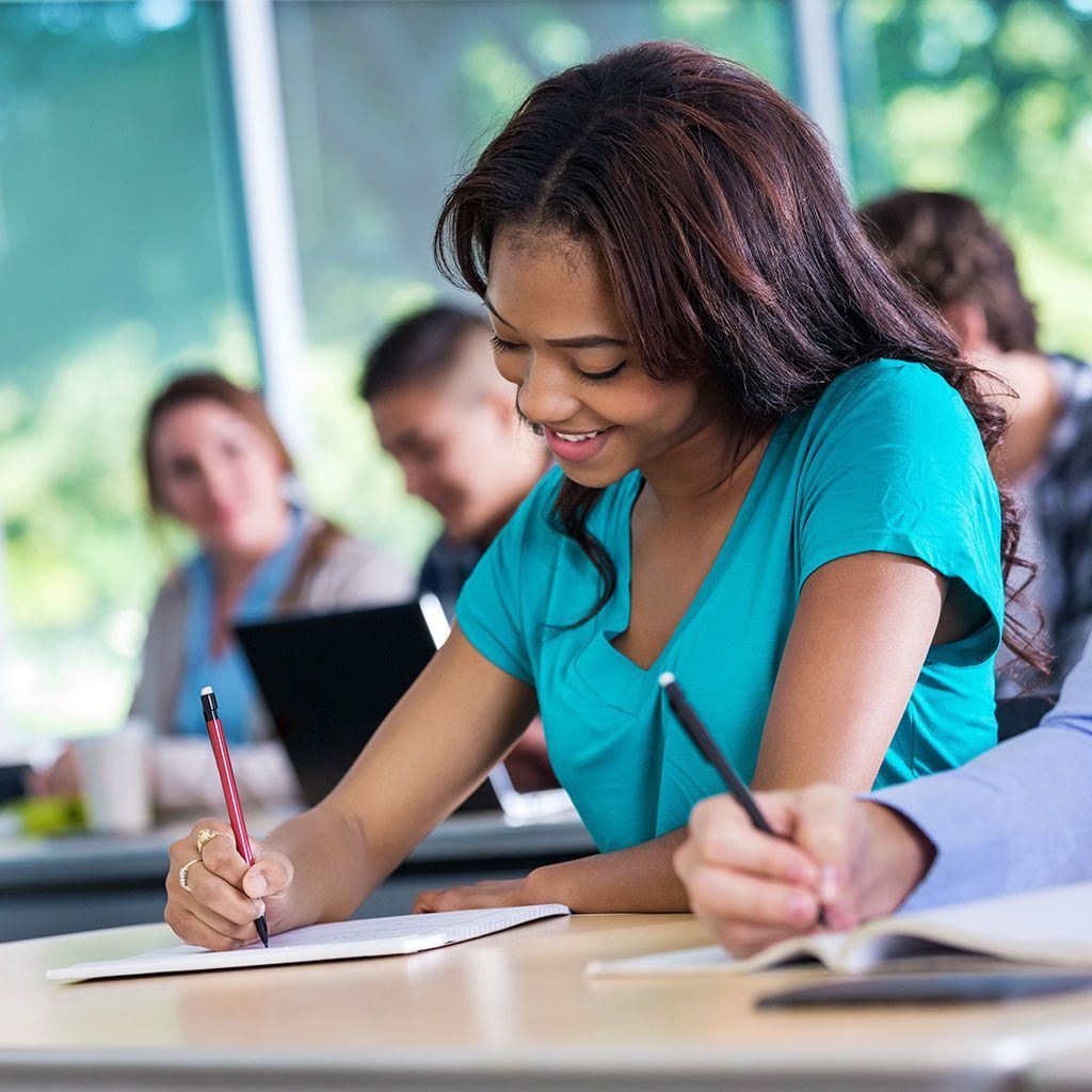 Una giovane donna sorridente scrive seduta a un banco in un'aula, in compagnia di altri studenti e alla luce del sole.