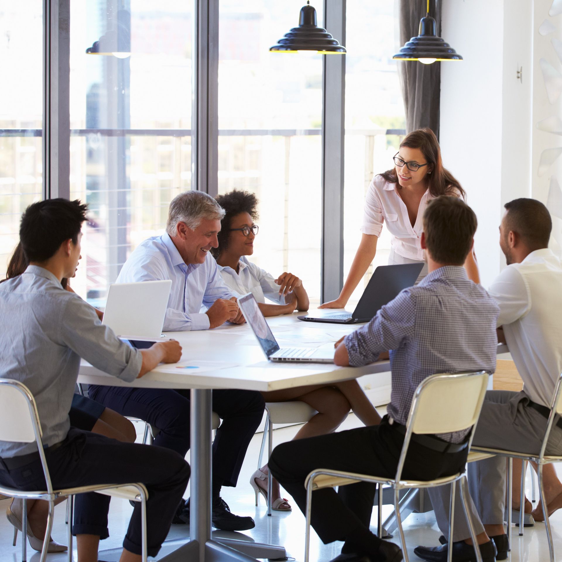 Un gruppo eterogeneo in abiti da lavoro è seduto a un tavolo, con i computer portatili aperti. Una donna è in piedi e si rivolge al gruppo.