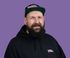 Man with beard wearing black hoodie and cap, purple background.