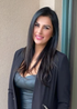 Woman in a black blazer and top smiles, leaning against a neutral wall.