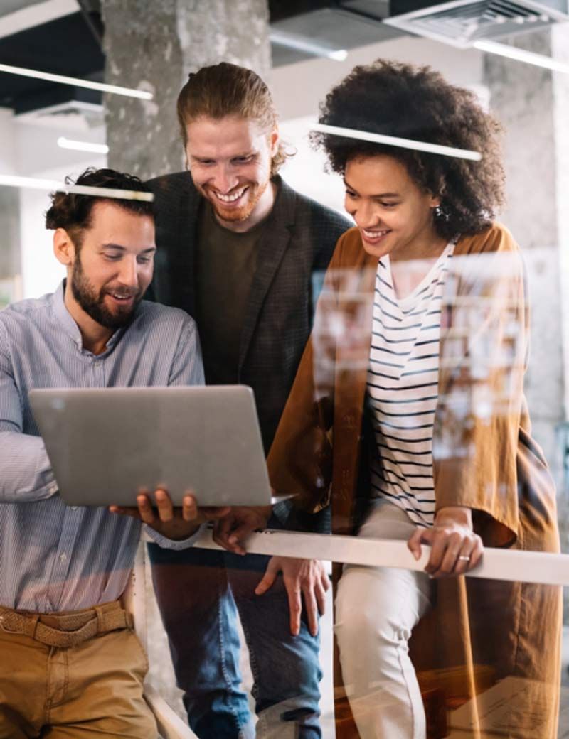 Three coworkers looking at a laptop in an office, smiling.