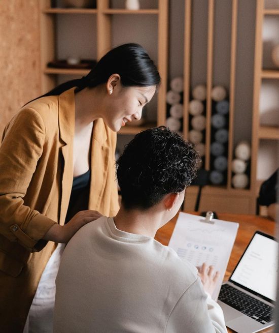 Woman in tan blazer smiles at a man at a desk; they review paperwork together.