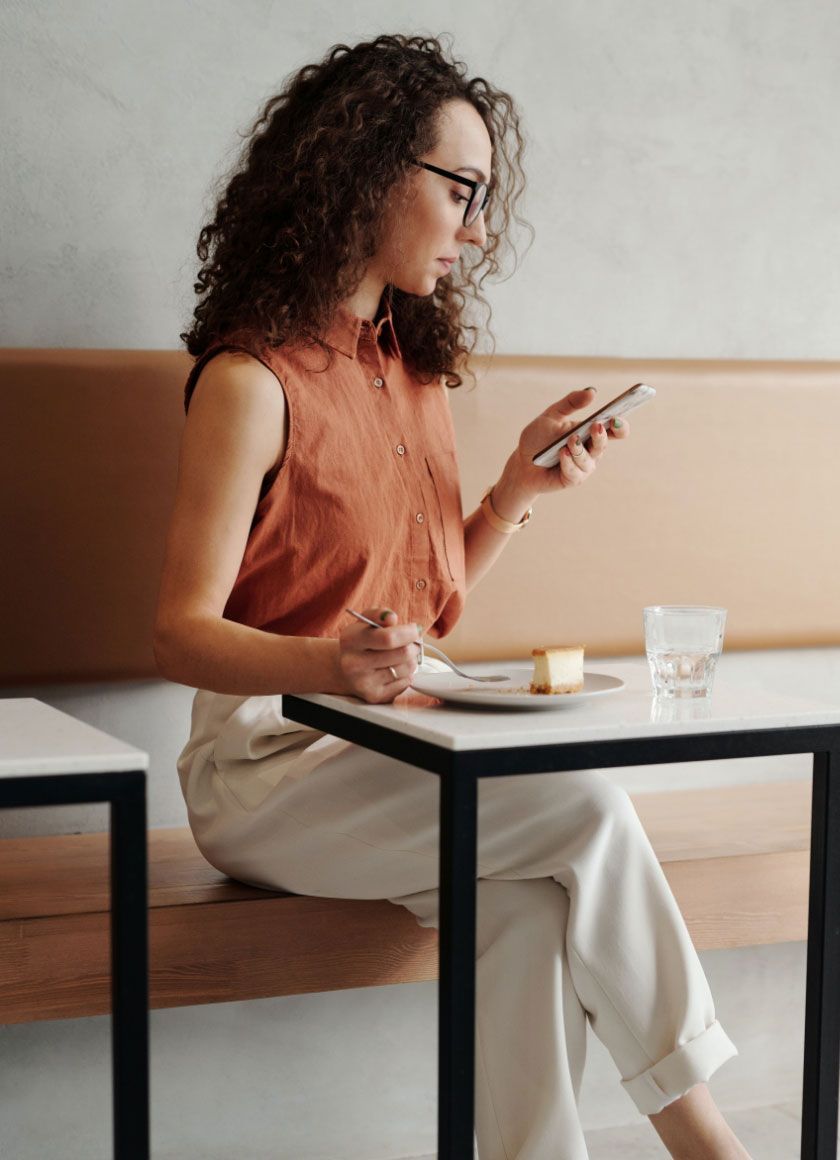 Woman with curly hair and glasses, sits at a table, looking at her phone while eating cake.