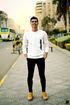Man in white sweater and black pants standing on a sidewalk, hands in pockets, smiling. City background.