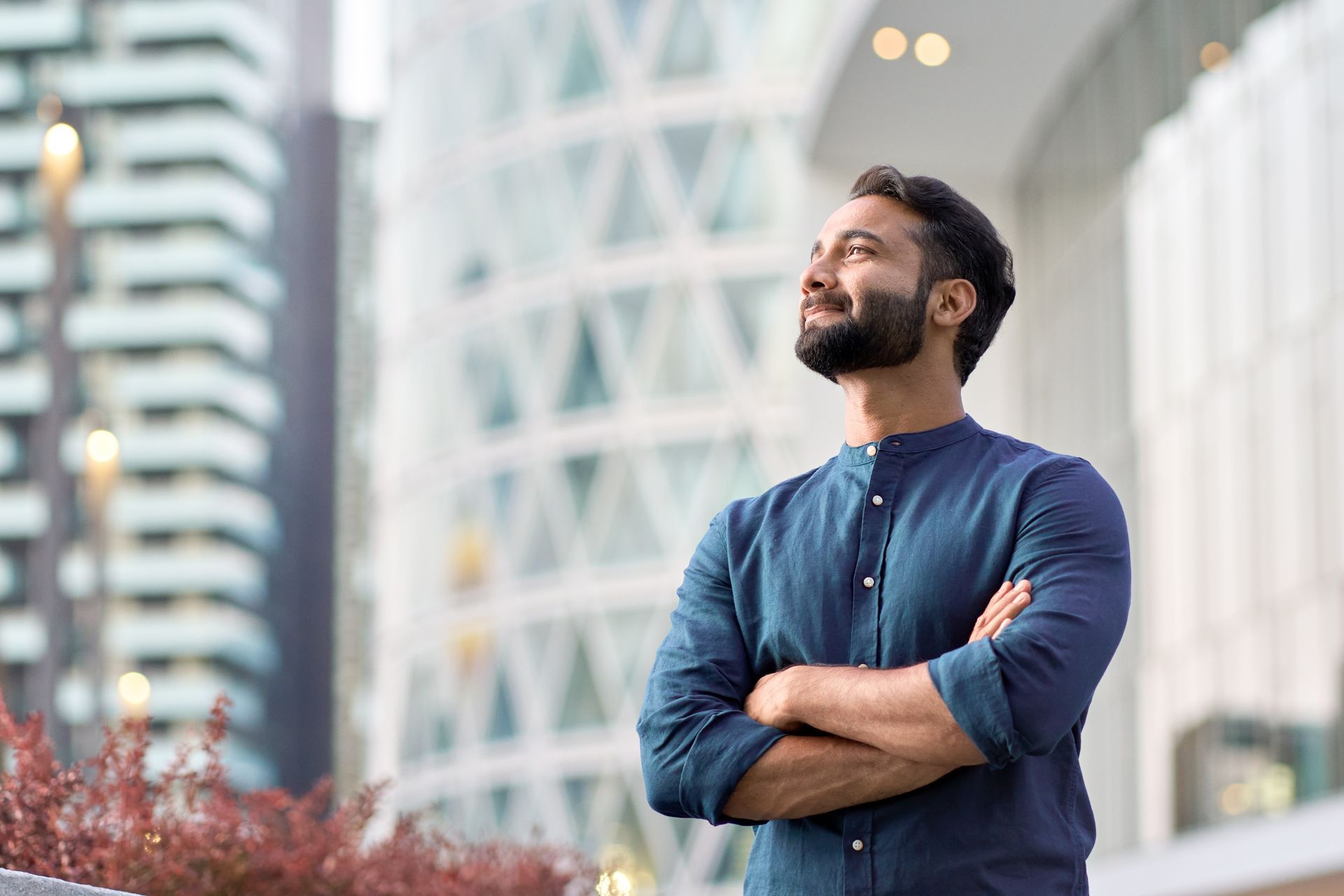 Man with arms crossed, looking upwards with a confident expression, standing in front of modern buildings.