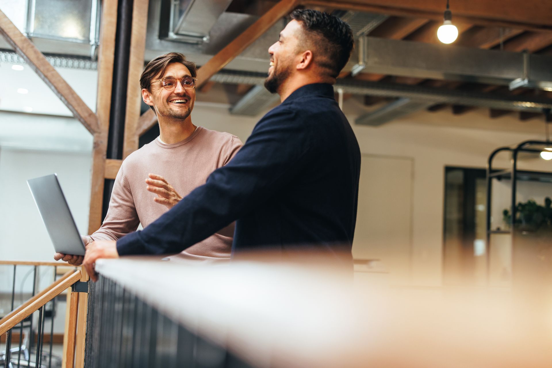Two men in modern office, smiling, one holding laptop, leaning on railing, wooden beams.