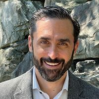 A man with a beard is smiling in front of a rock wall.