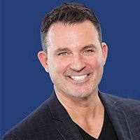 A man in a suit and black shirt is smiling in front of a blue background.