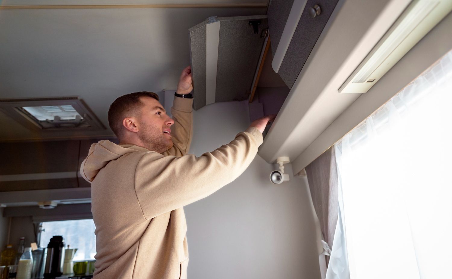 A person in a beige hoodie reaches up to open a grey cabinet door inside a camper or RV.