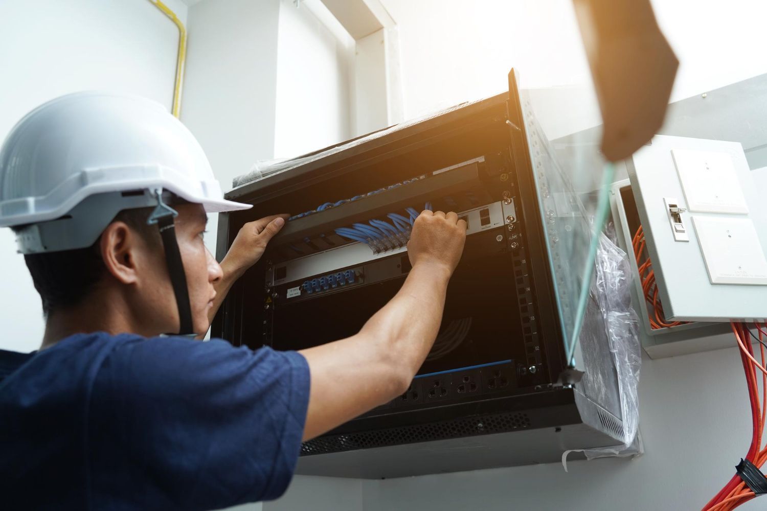 A technician in a hard hat installs network equipment into a wall-mounted server rack.