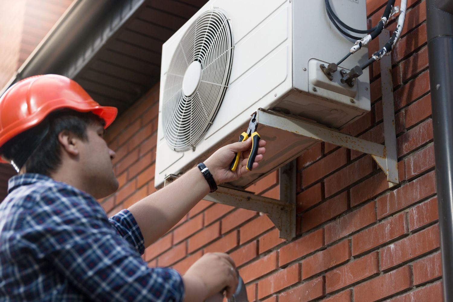 A technician in a hard hat uses pliers to work on an air conditioning unit mounted on a brick exterior wall.