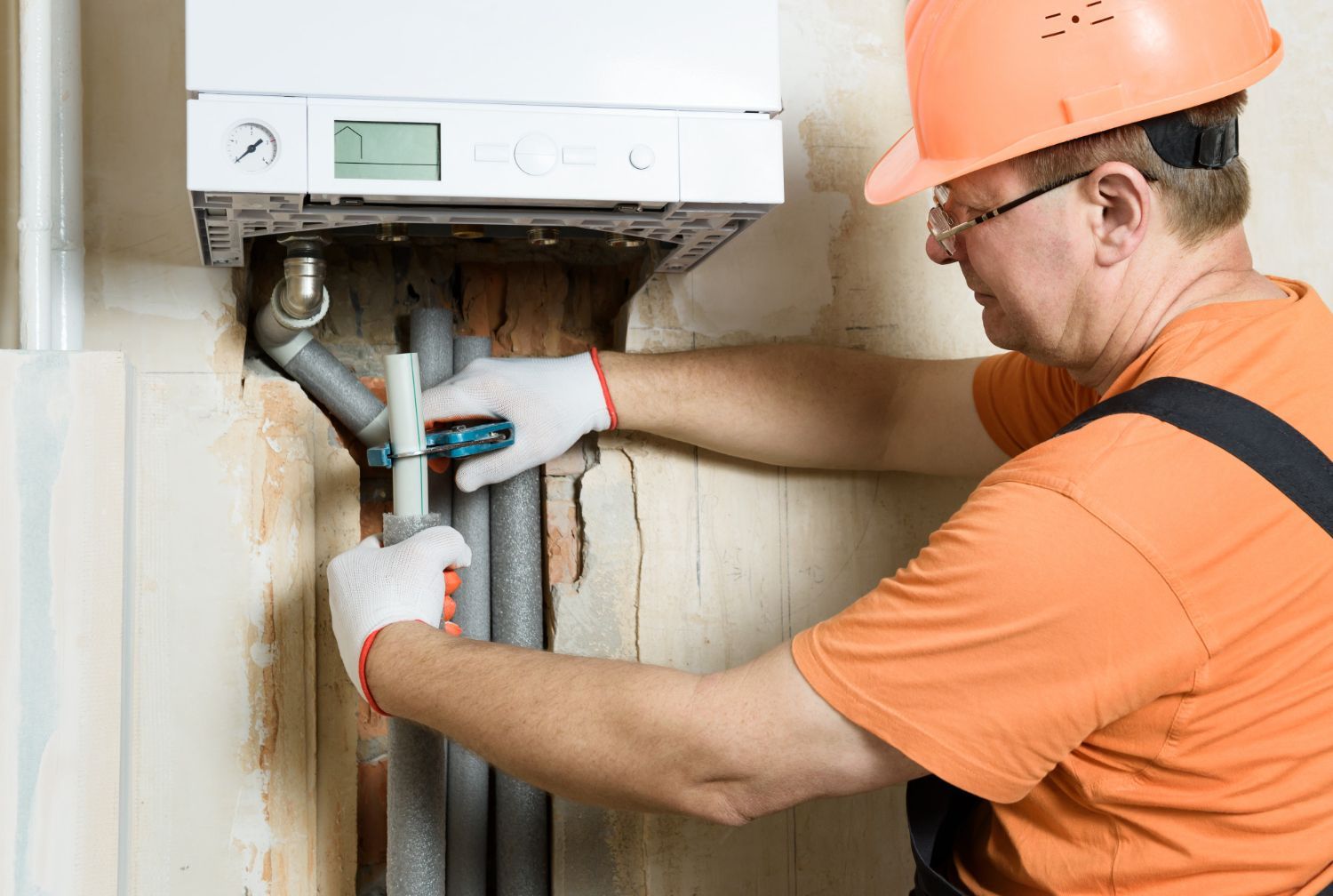 A technician in an orange shirt and hard hat wearing work gloves installs pipes beneath a wall-mounted gas boiler.