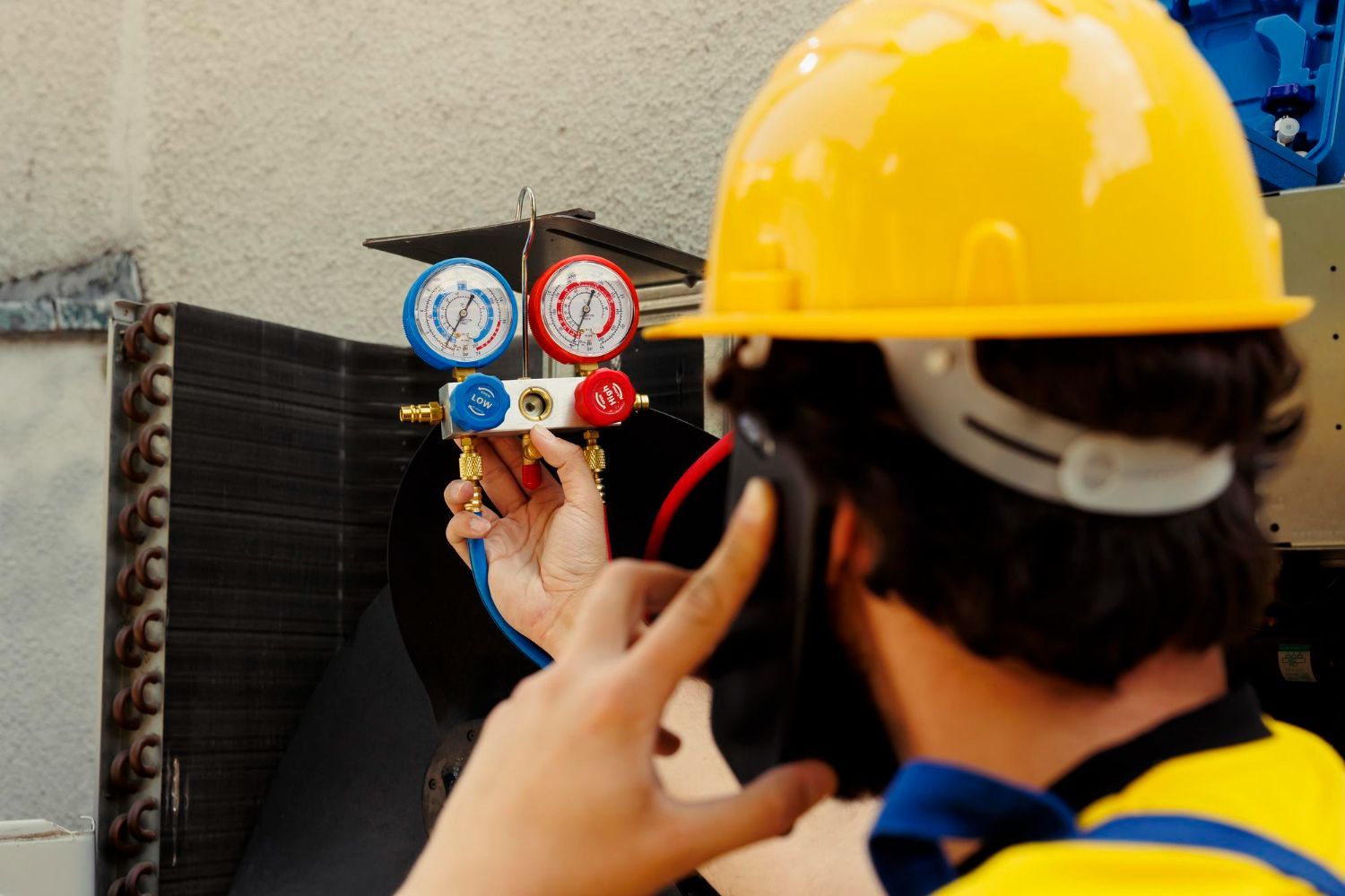 A worker in a yellow hard hat checks HVAC manifold gauges while talking on a smartphone.