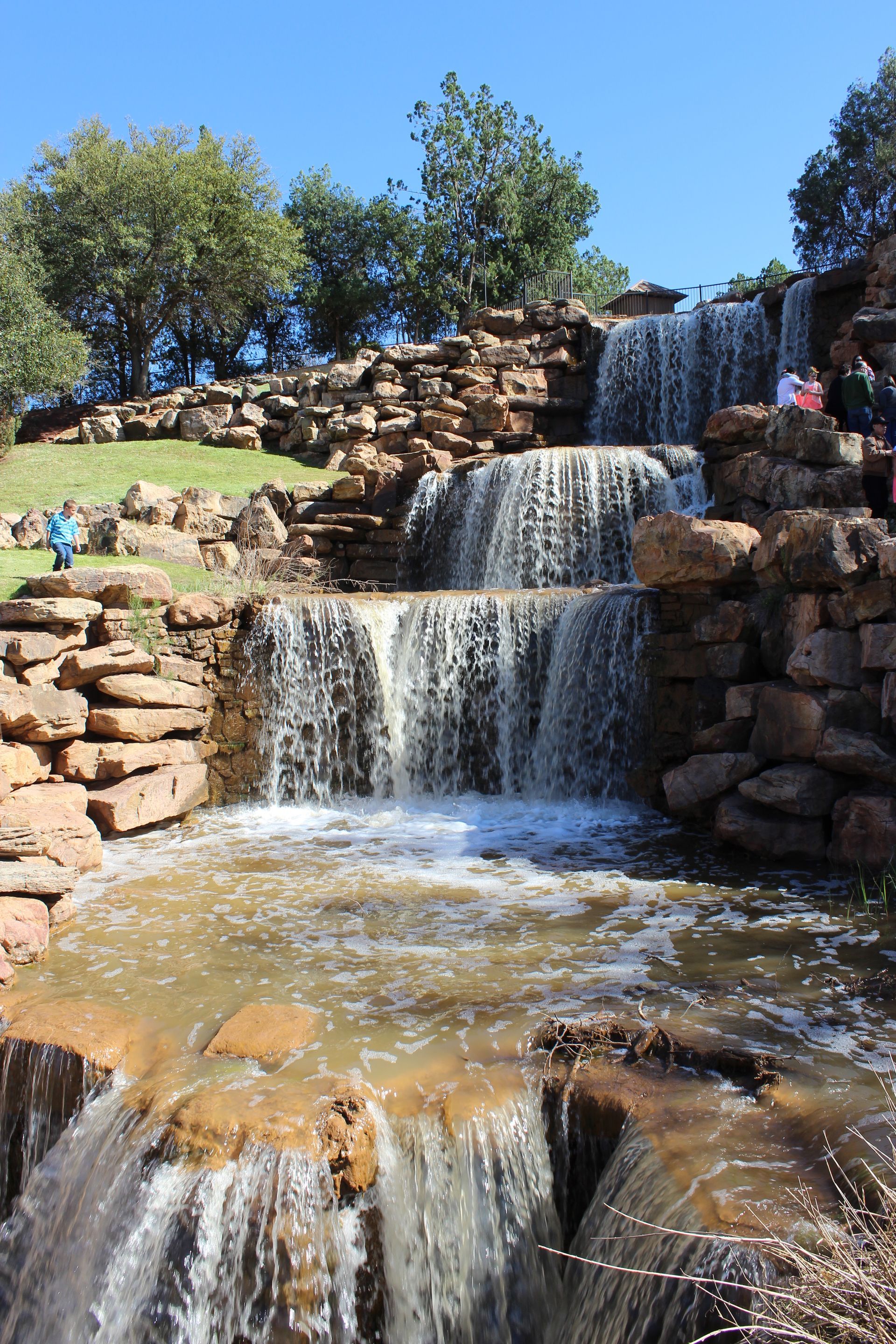 A multi-tiered waterfall cascades over brown rock formations into a pool under a clear blue sky.