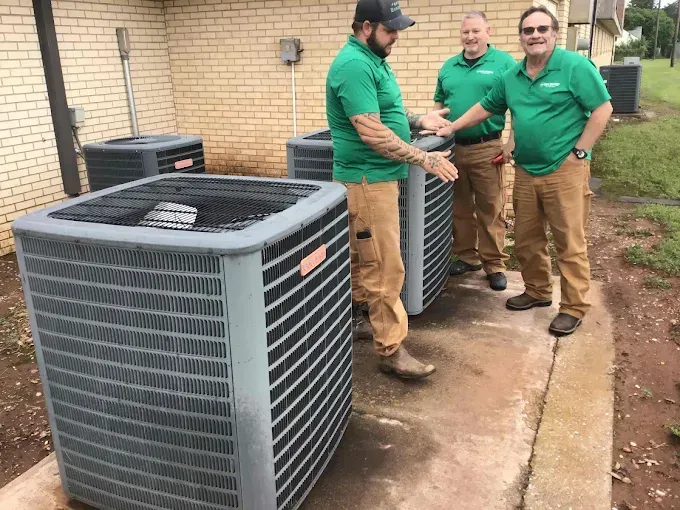 Three technicians in matching green shirts and tan pants stand beside large outdoor air conditioning units at a building.
