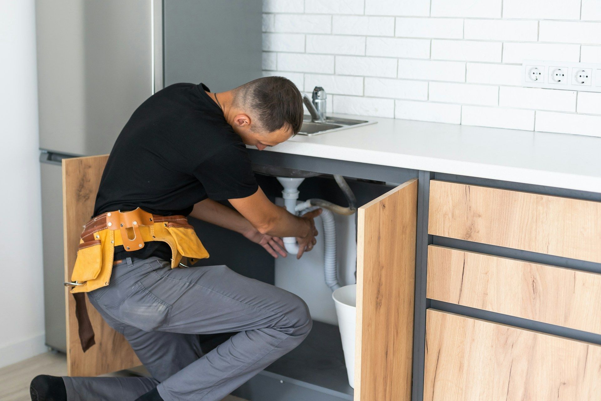 A plumber in a black shirt and tool belt repairs pipes under a kitchen sink with white subway tile in the background.