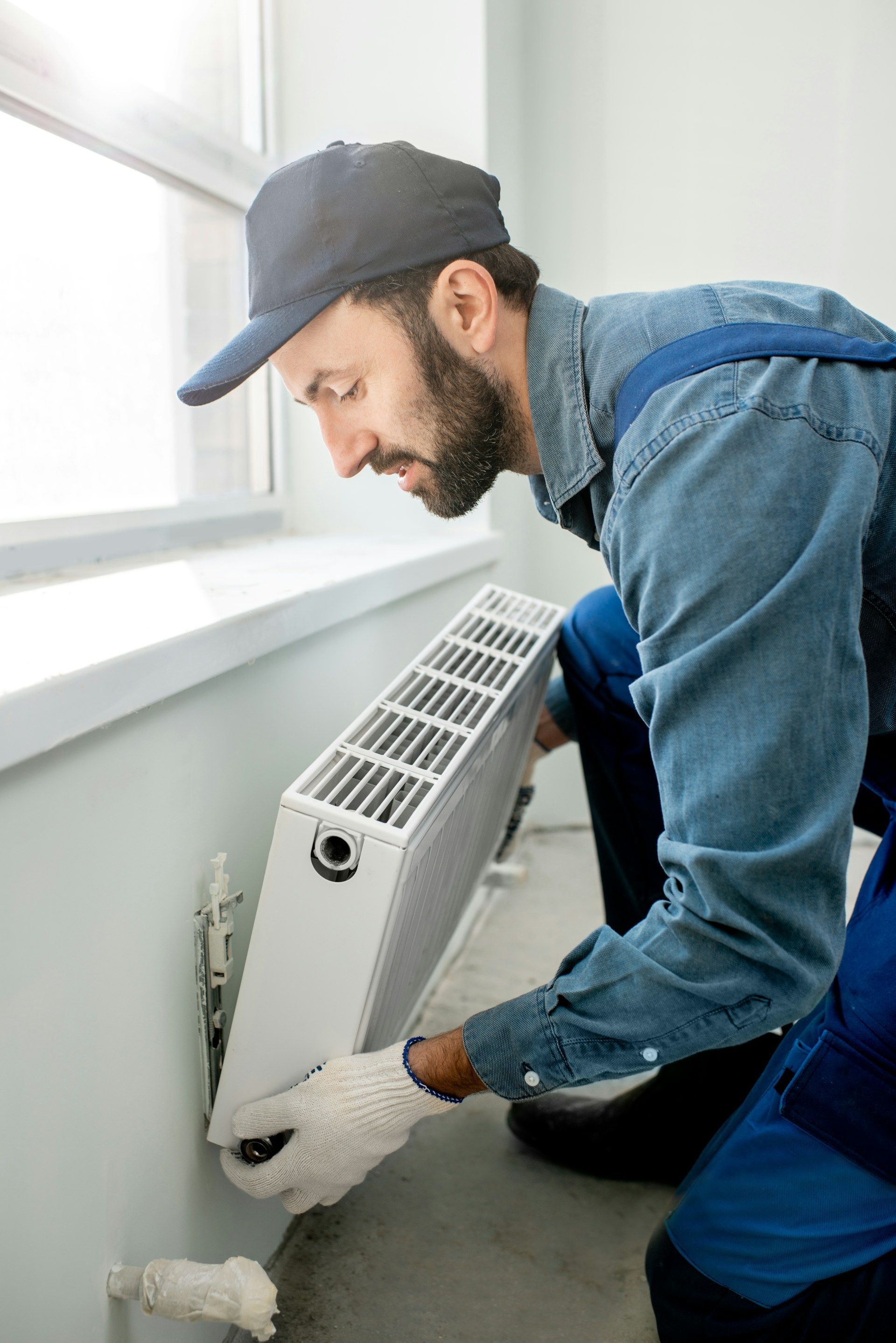 A worker in a cap and denim jumpsuit installs a white radiator on a wall beneath a window.