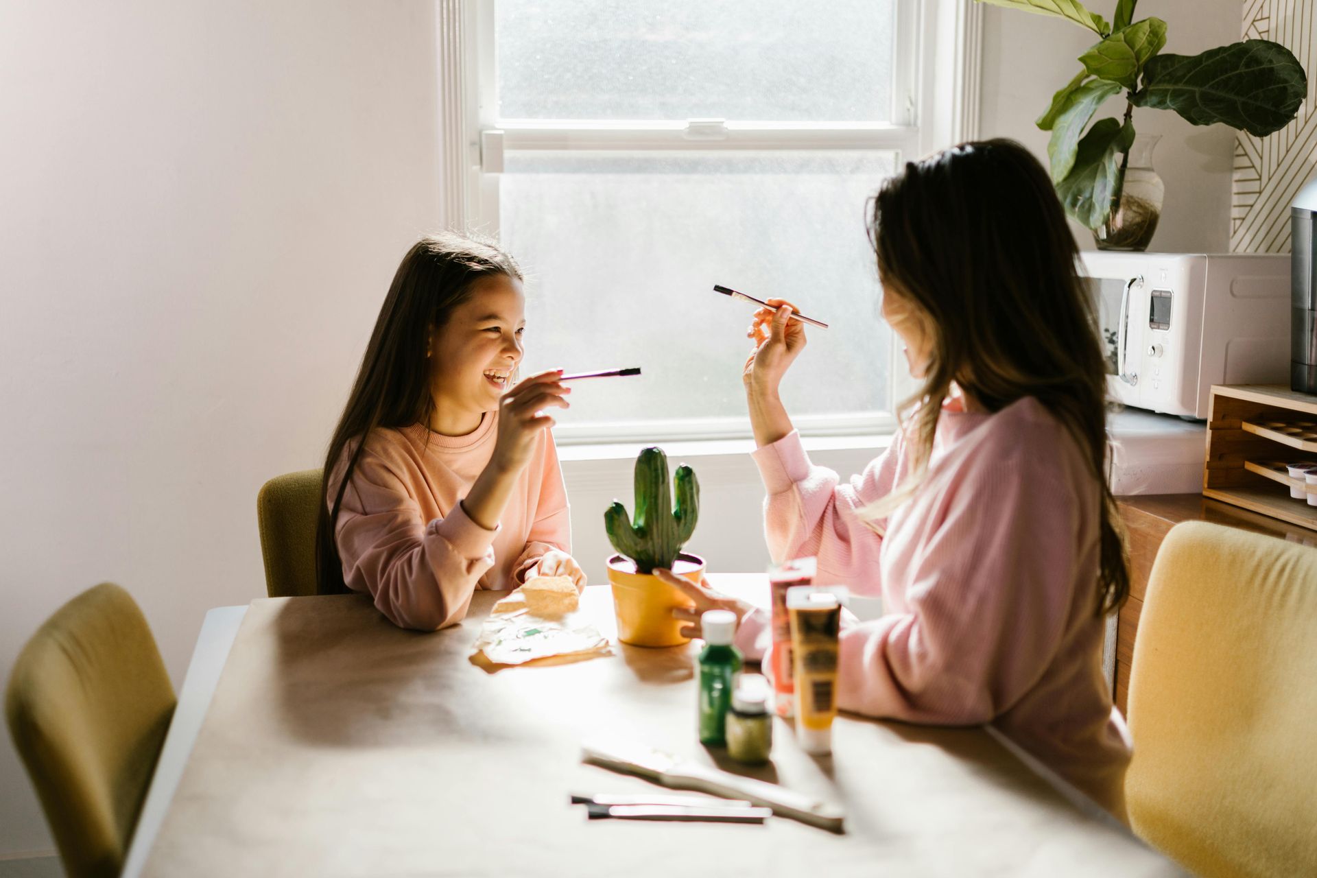 A person and a child wearing pink sweaters sit at a table painting a small cactus decoration in a sunlit room.