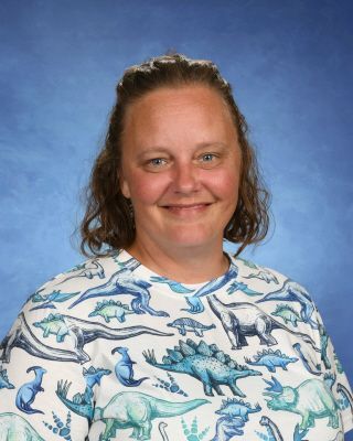 A woman wearing a shirt with dinosaurs on it is smiling for the camera.