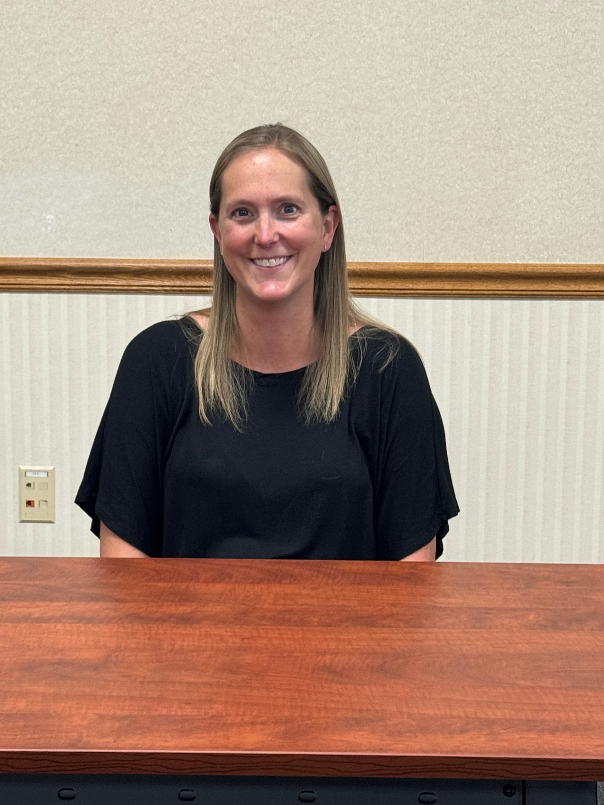 A woman in a black shirt is sitting at a wooden desk and smiling.This is Christa Heckman.