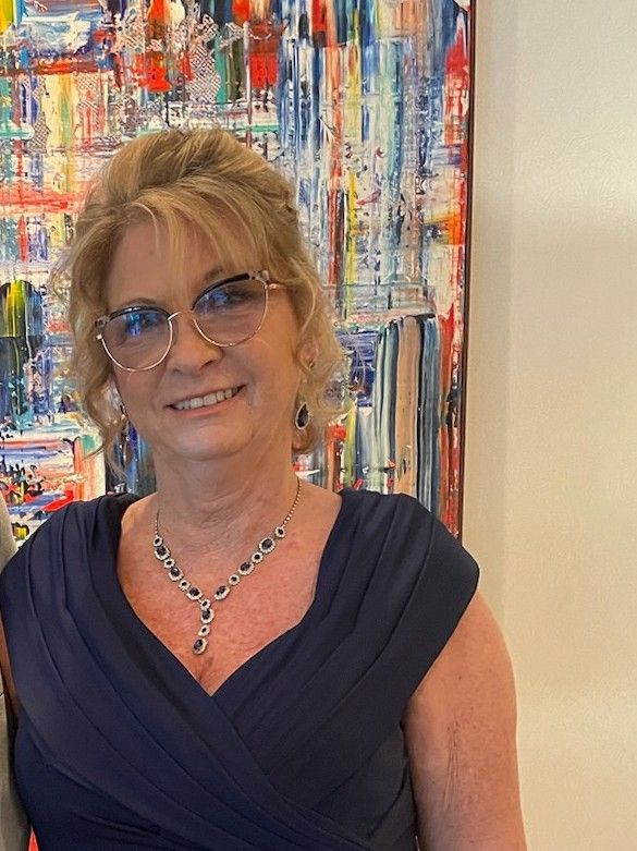 A woman wearing glasses and a blue dress is standing in front of her classroom. This is Cynthia Bartholomew.