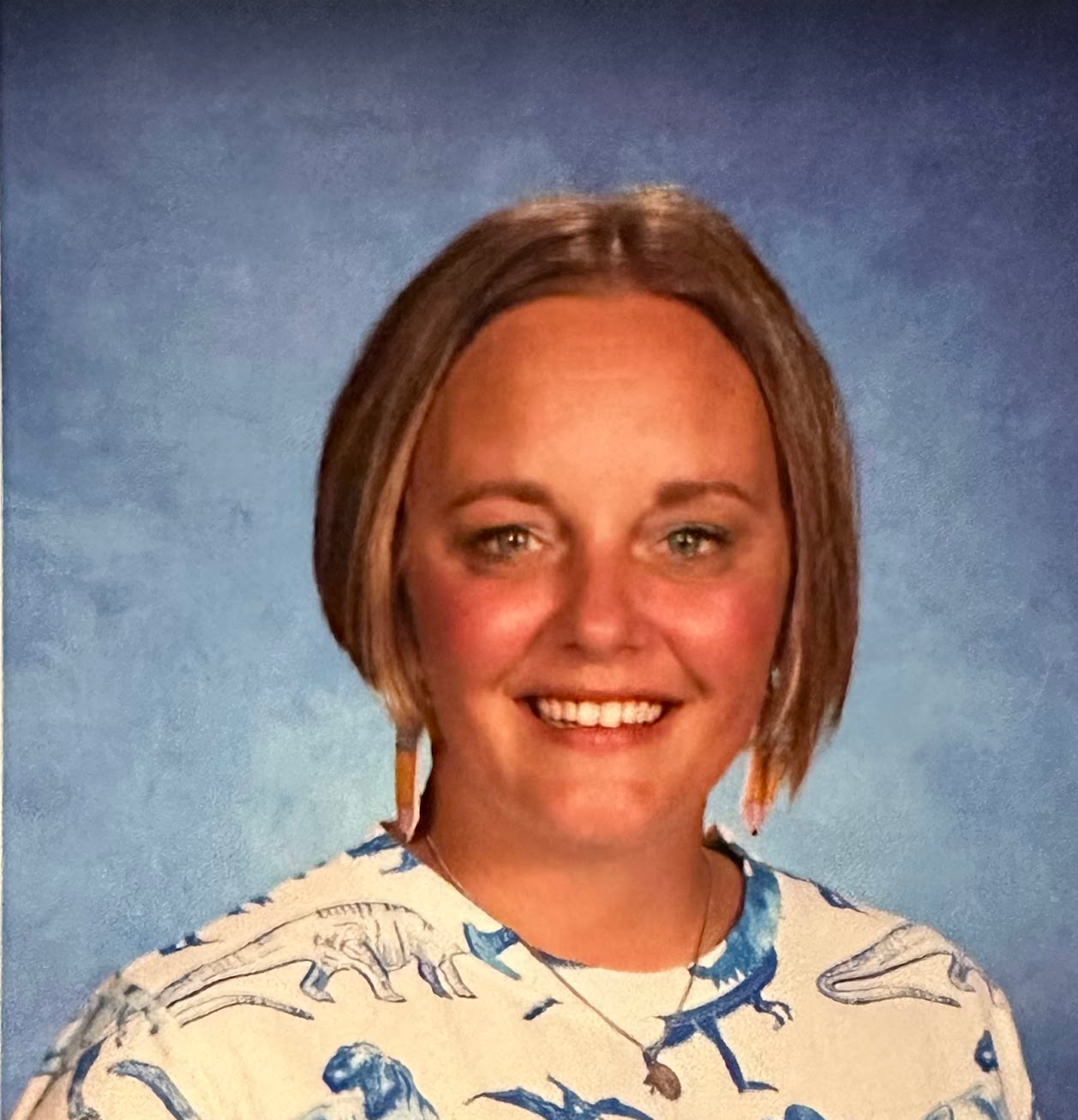 A woman wearing a shirt with dinosaurs on it is smiling for the camera.