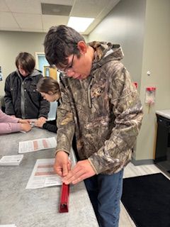 A boy in a camo jacket is cutting a piece of paper with a ruler.