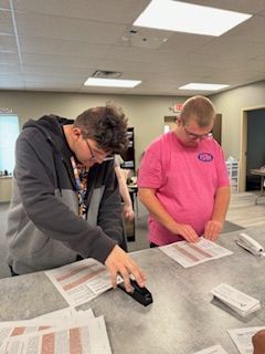 Two men are sitting at a table working on papers . one of the men is wearing a pink shirt.