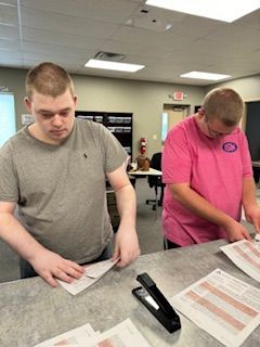 Two men are working on a table with papers and a stapler.