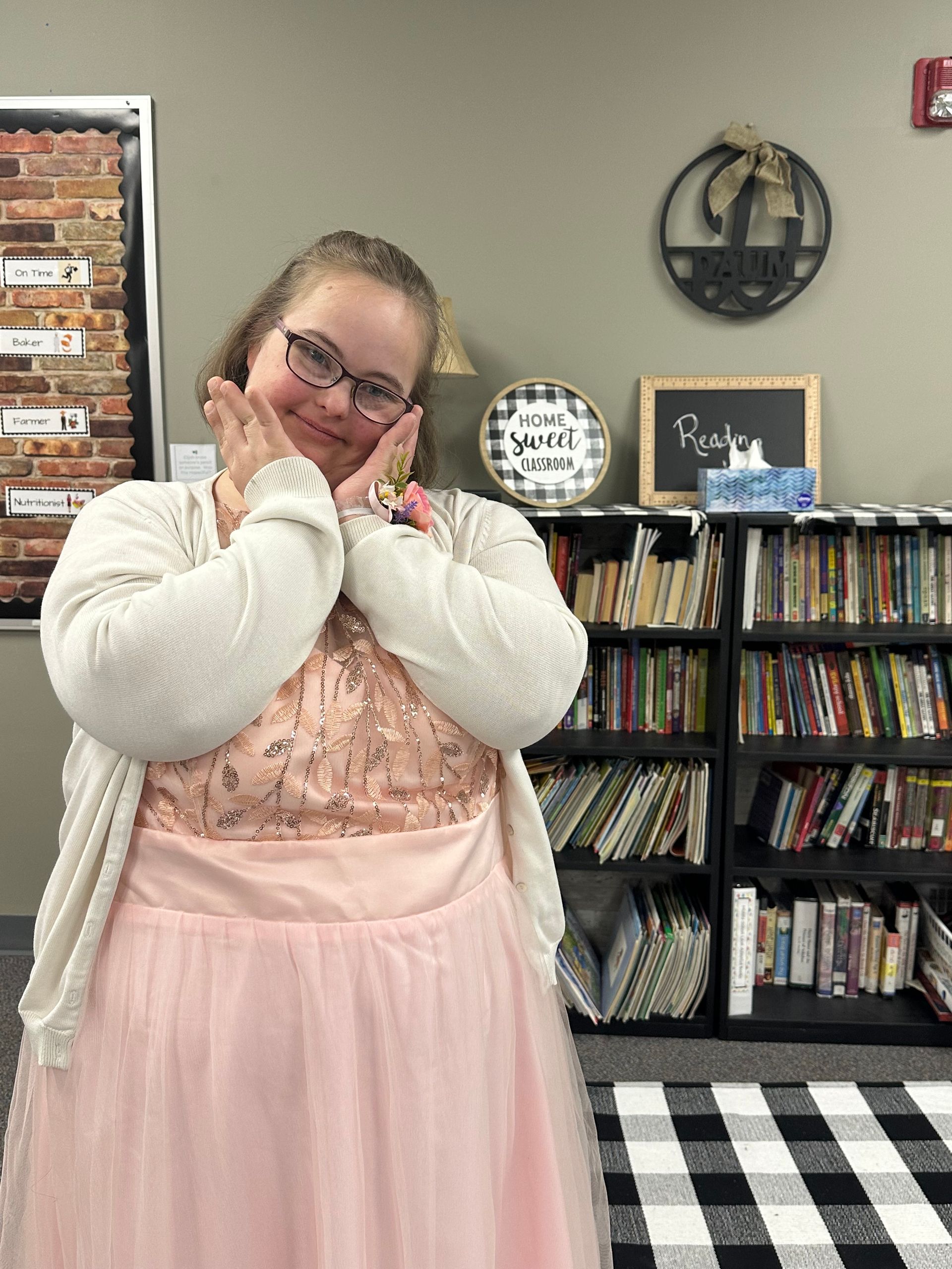 A woman in a pink dress and white cardigan is standing in front of a bookshelf.