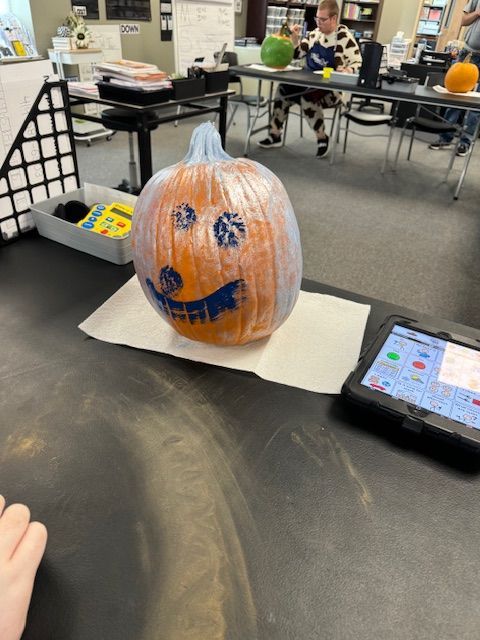 A pumpkin with a face painted on it sits on a table next to a tablet