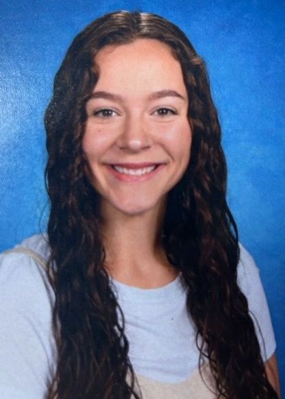A woman with long brown hair is smiling in front of a blue background.