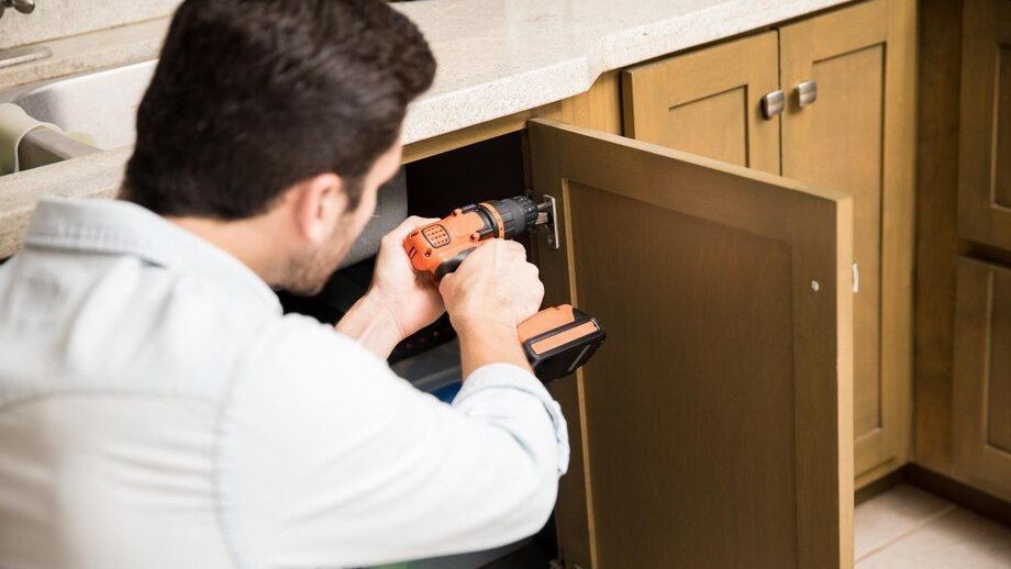 A man is using a drill to fix a cabinet in a kitchen.