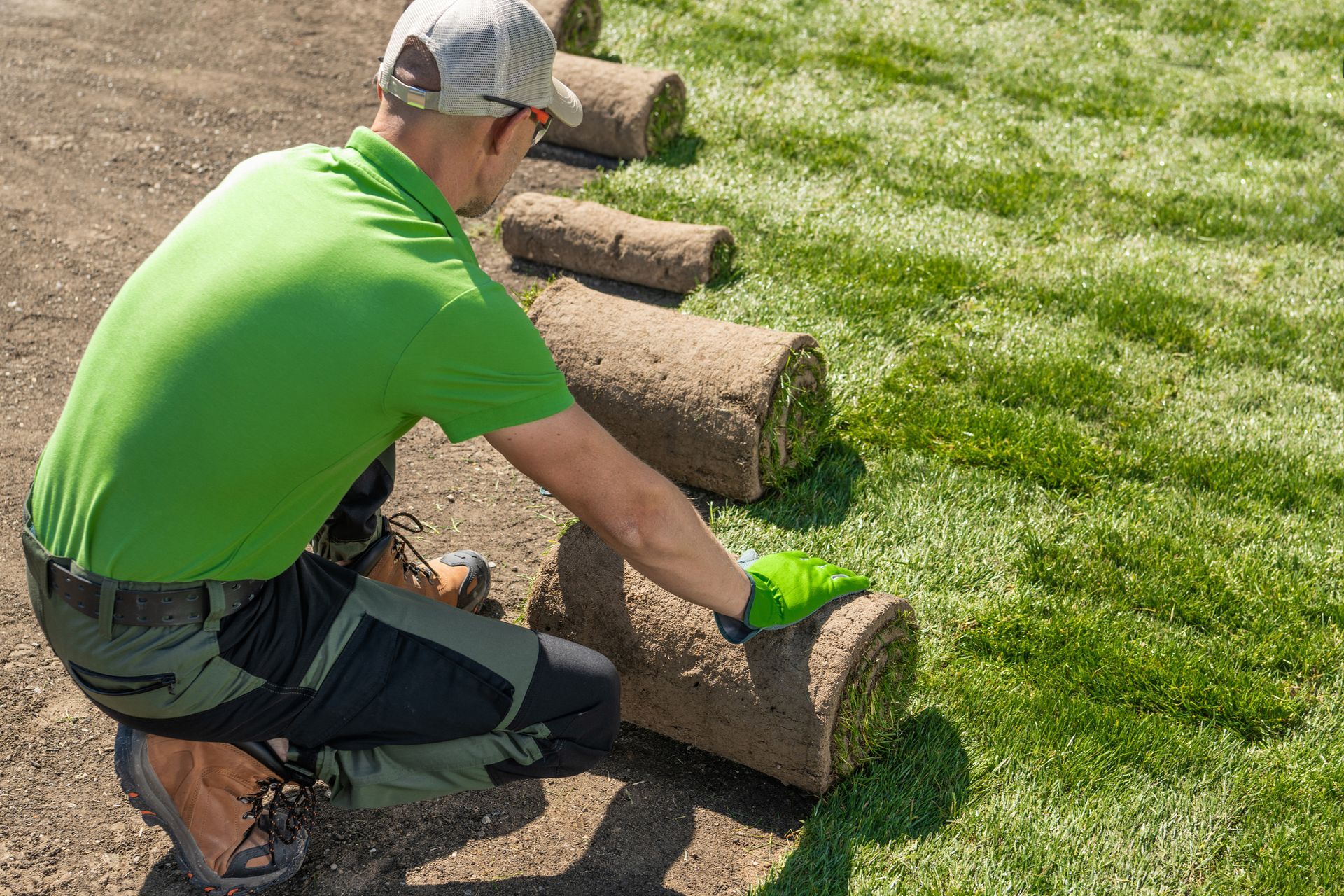 A man in a green shirt is kneeling down and rolling a roll of turf.