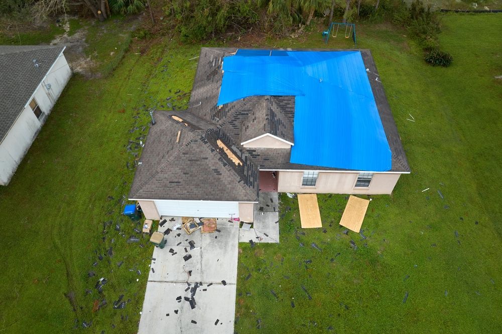 Aerial view of a house with significant roof damage. Blue tarp covers part of the roof. Debris and plywood are scattered around.