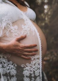 A pregnant woman in a white lace dress is holding her belly.