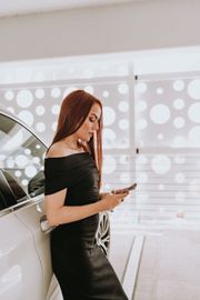 A woman in a black dress is leaning against a white car and looking at her phone.