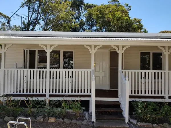 White cottage with porch, railing, front door, and steps, surrounded by greenery.