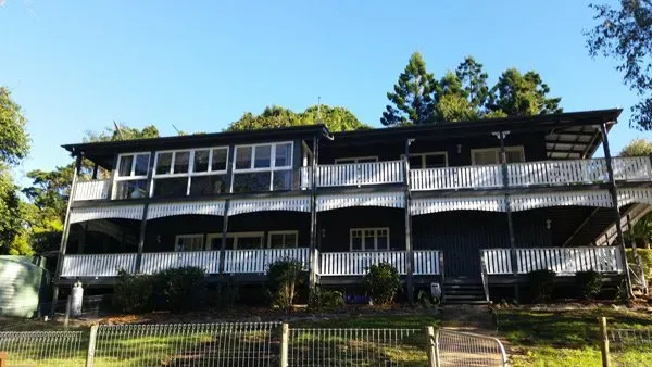 Two-story dark house with white railings and windows, set among trees under a blue sky.
