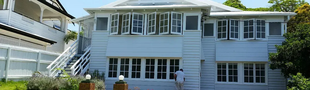 White house with multiple windows and open shutters. A person is painting the exterior.