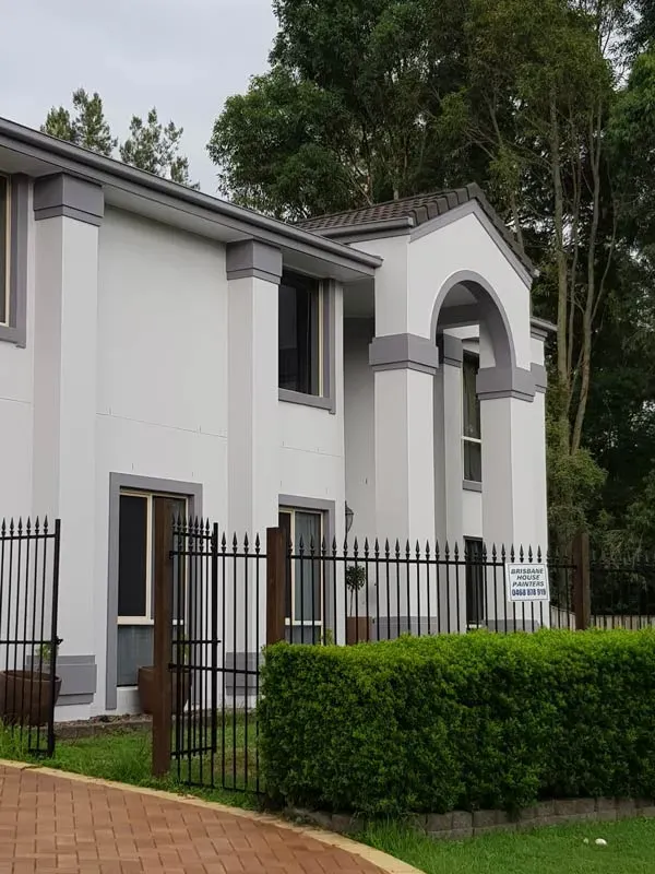 Two-story building with gray trim and white walls behind a black fence and green hedge.