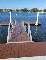 Wooden dock extending over water with metal ramp and white posts. Blue sky, boats in background.