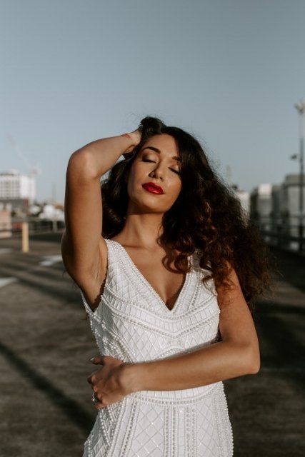 A woman in a white dress is standing in a parking lot with her eyes closed.