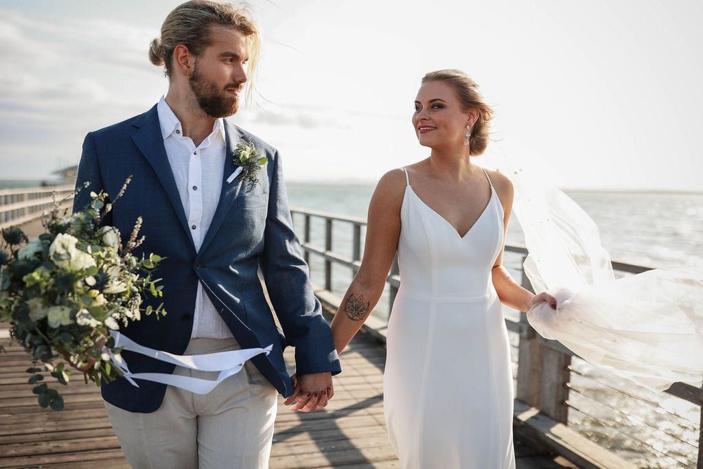 A bride and groom are walking on a pier holding hands.
